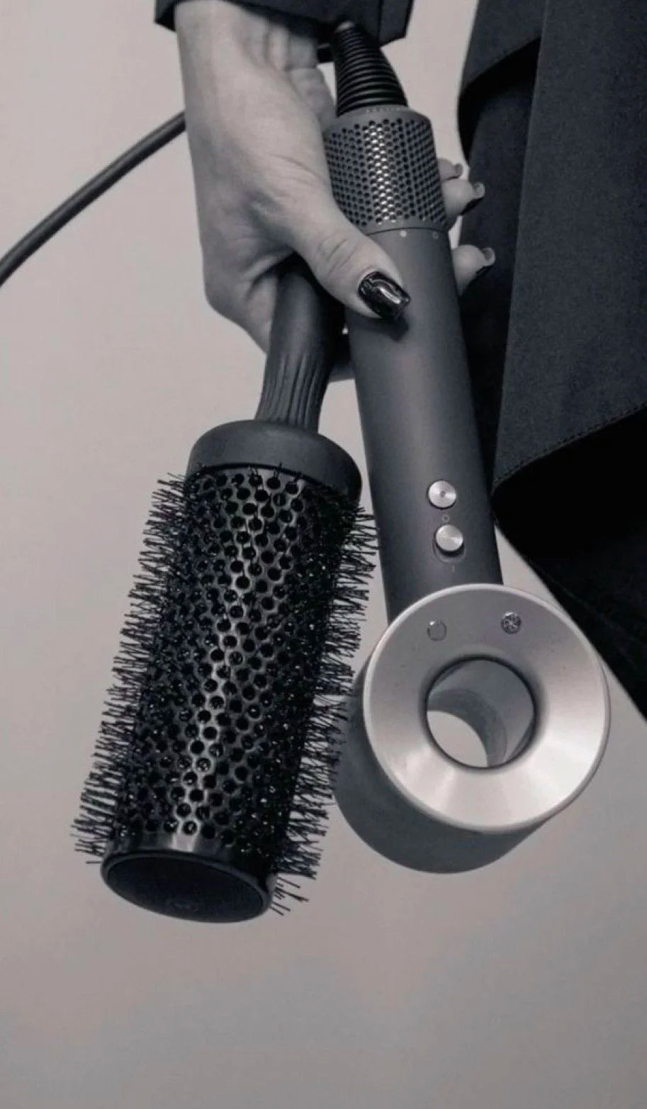Close-up of a person holding a black round hairbrush and a gray cordless hair dryer with silver buttons and nozzle, against a plain background. at Eve Salon, Saint Louis MO