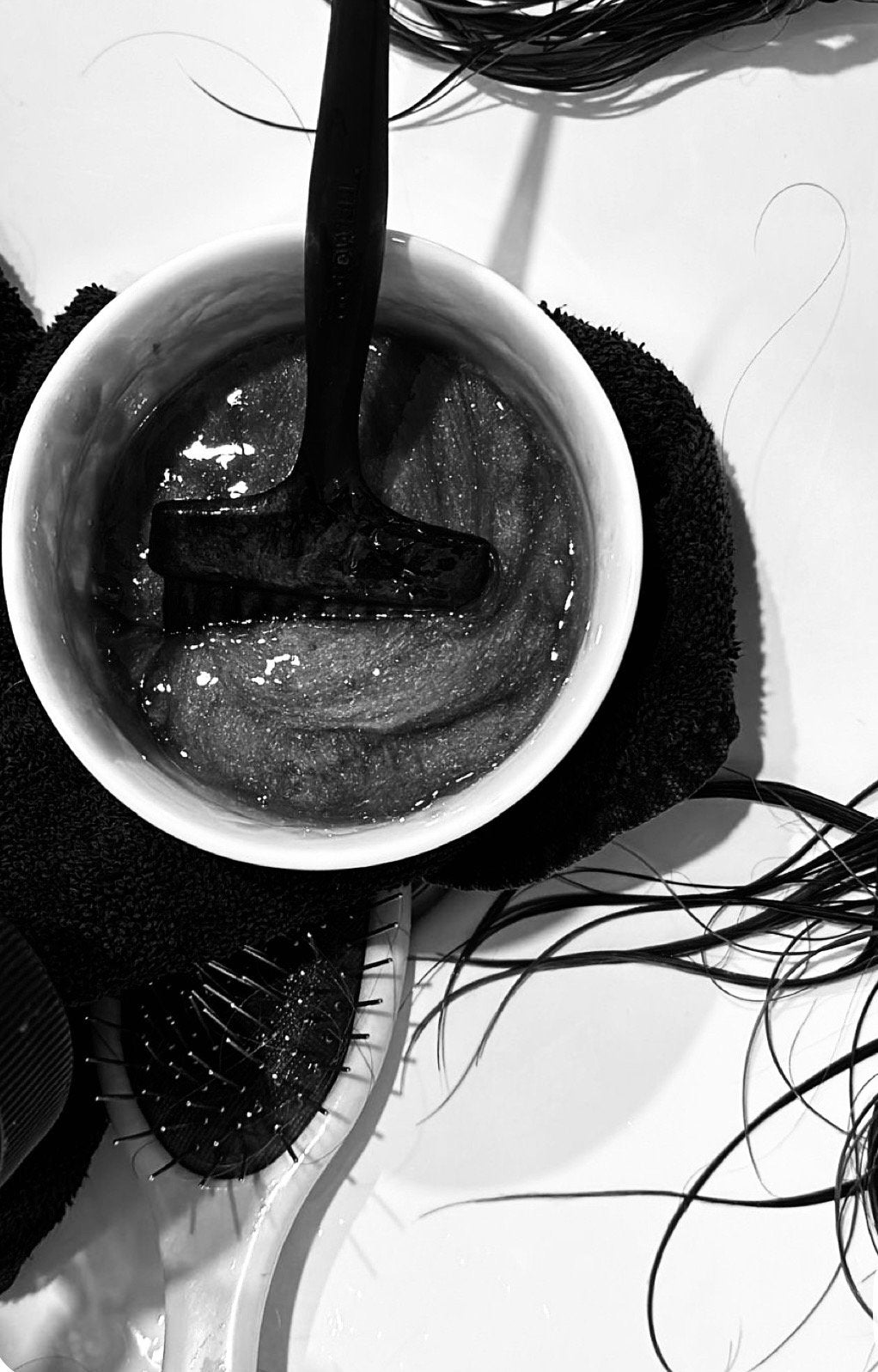 A black and white photo of a hairbrush with black bristles, a cup with hair dye and a black applicator brush inside, surrounded by hair strands, and a towel underneath the cup. at Eve Salon, Saint Louis MO